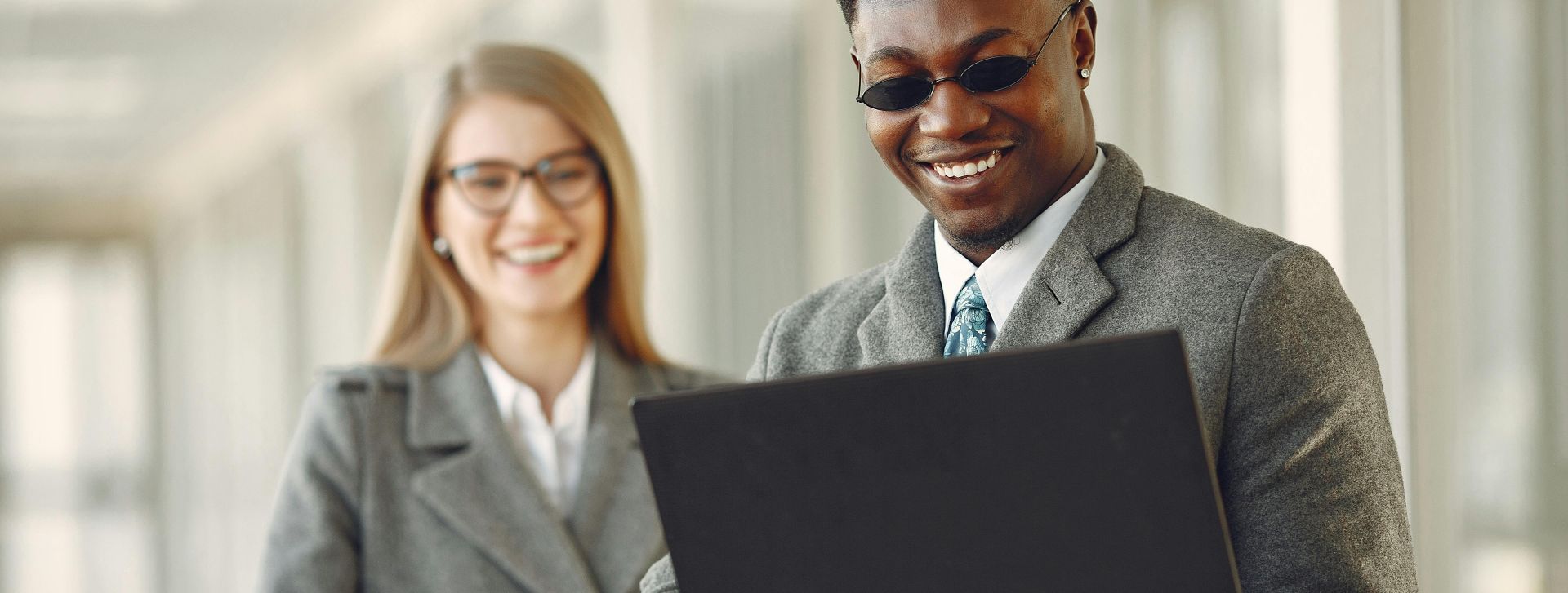 Positive smiling stylish diverse business people in grey formal outfits  and trendy eyeglasses browsing netbook while standing in contemporary workspace in daylight