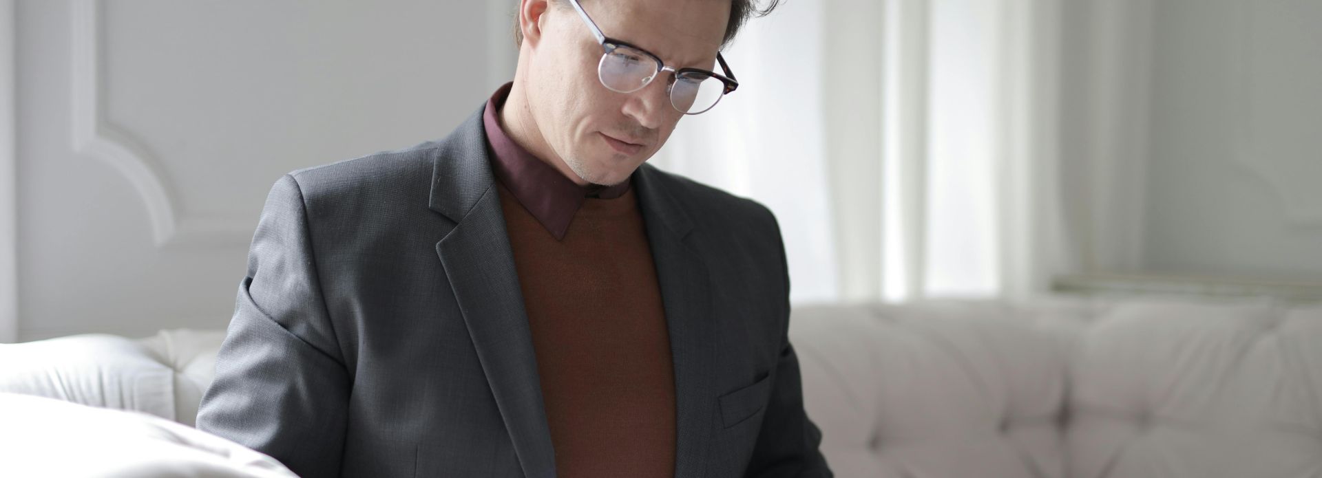 Elegant adult man in jacket and glasses looking through documents while sitting on white sofa in luxury room